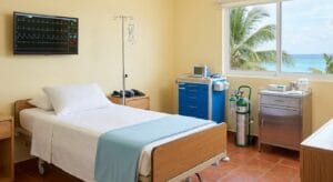 Medical treatment room at Iboga Wellness Institute's Cozumel clinic equipped with cardiac monitoring equipment, IV pole, patient bed, and emergency medical supplies in a bright, naturally-lit space