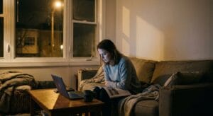 A woman sits on a sofa late at night in a quiet living room, reading printed pages while a laptop glows on the coffee table beside an untouched cup of tea.