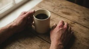 Close-up of weathered hands gripping a white mug on a wooden table in morning light, conveying the tension and quiet struggle of early recovery